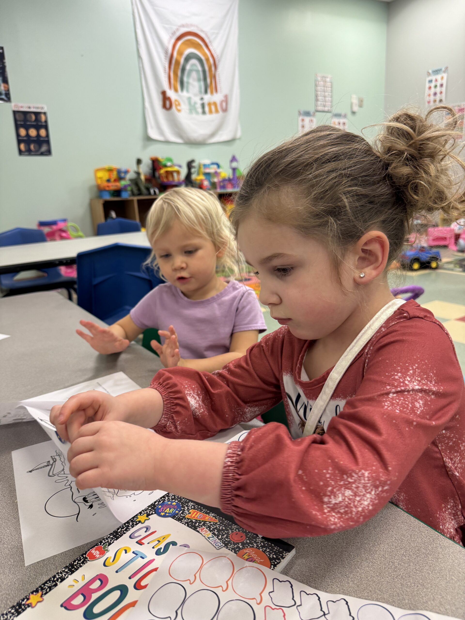 Two children in a well-lit room at a desk with coloring sheets and stickers.